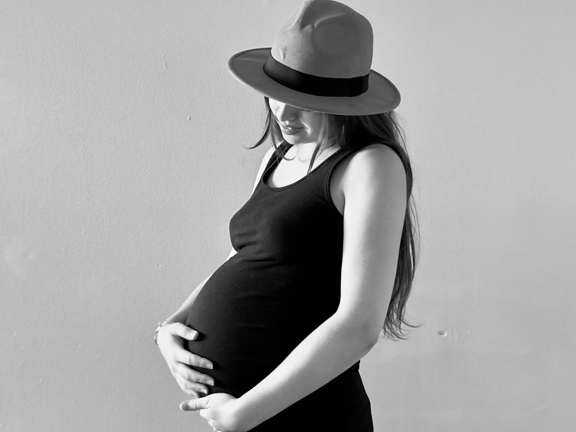 Black and white portrait of a pregnant woman in a hat gently holding her baby bump – symbolizing the expectations and emotions before becoming a new mom